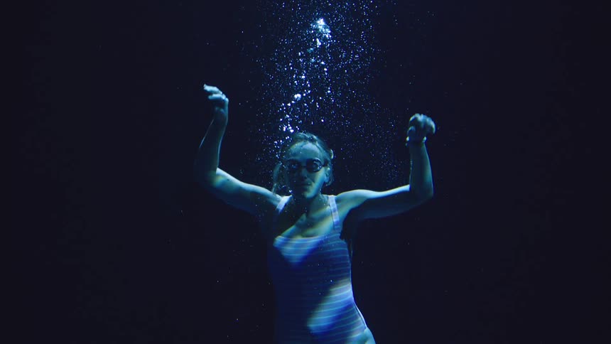 A woman in goggles floats underwater as bubbles stream upward through blue light