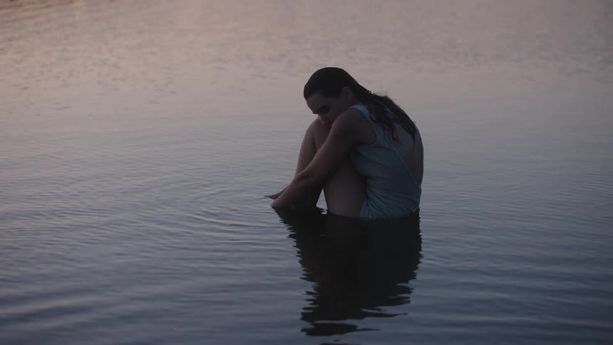 A young woman sits alone in shallow still water at sunset wearing a swimsuit the golden light reflects softly across the surface as she gazes downward