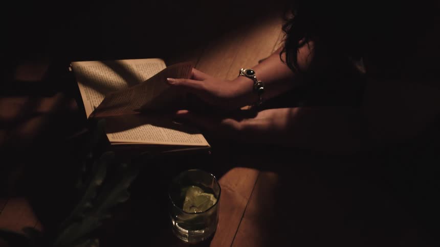 The hands of a woman wearing a silver bracelet holds open a small book next to a glass of iced drink on a wooden floor with a shaft of light illuminating the pages in a dark room