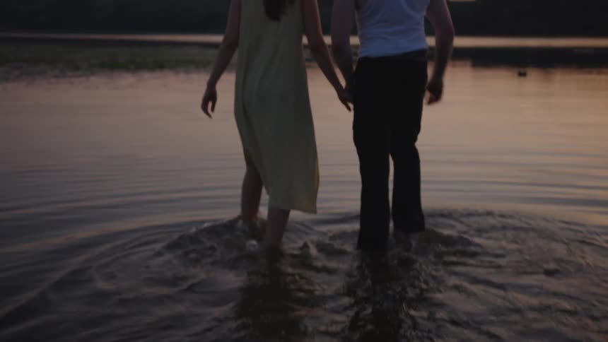 A shot of a couples legs and lower torsos shows them standing in shallow water at sunset holding hands and wading through the liquid