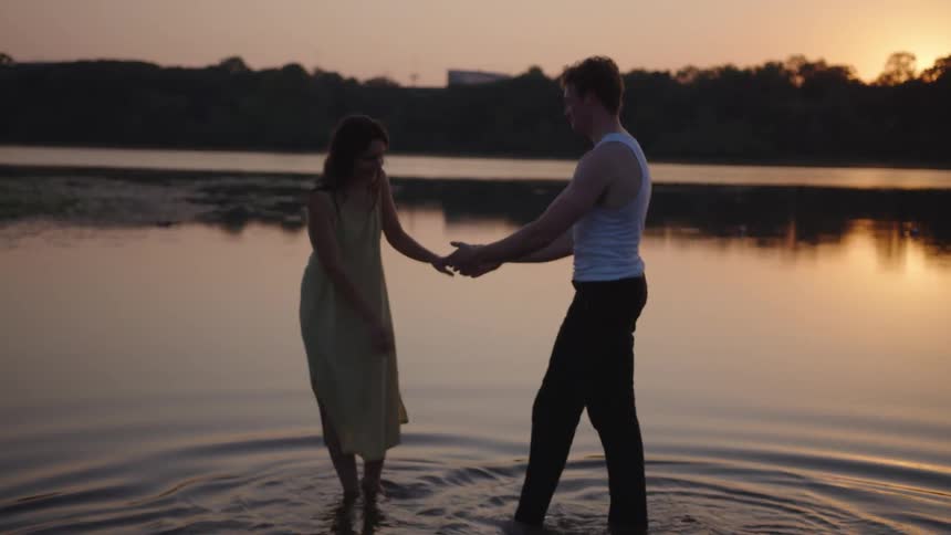 A man and a woman hold hands and dance while standing ankledeep in a lake at sunset creating gentle ripples