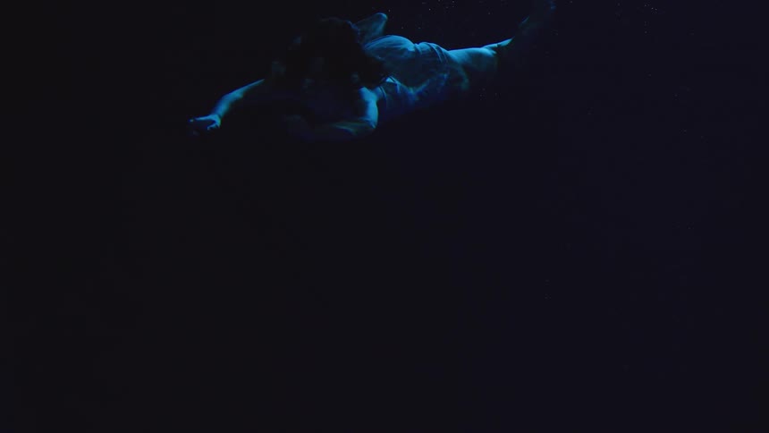 A woman in a pale blue outfit is suspended in dark blue water with her arms spread wide and her body slowly sinking into the deep gloom