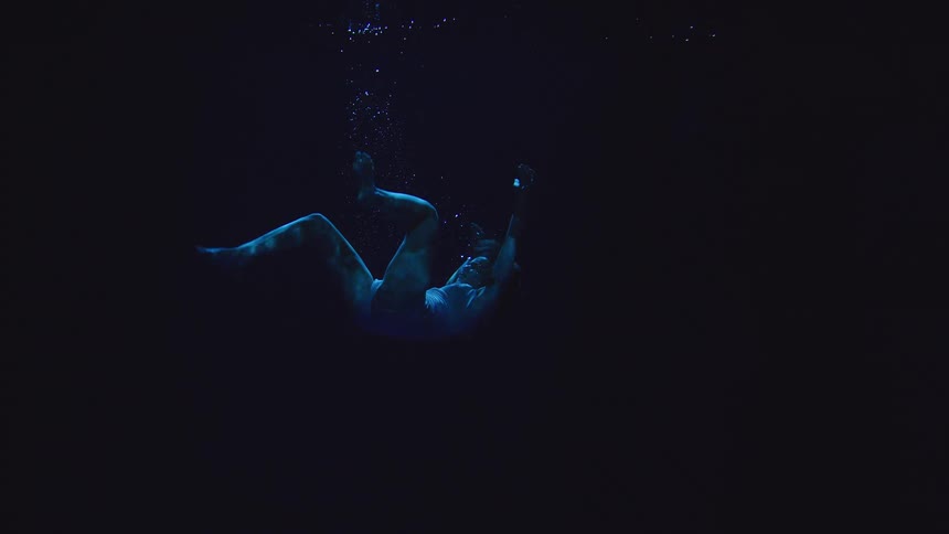 A woman in a pale blue swimsuit is seen underwater floating and slowly sinking into the dark blue void with a few air bubbles rising above her