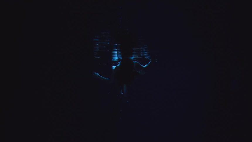 A person swims underwater in darkness with arms outstretched silhouetted beneath faint blue reflections on the surface