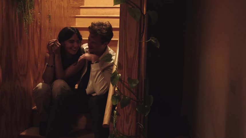A man and a woman smile and laugh while they sit on a wooden staircase in warm light the woman covers her face from the rain