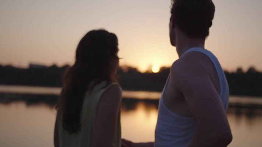A young couple embraces by the riverbank at sunset their silhouettes glowing softly in the golden light as the water reflects the fading sky