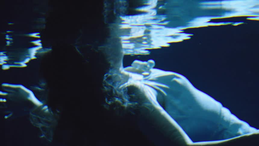 A young woman wearing a white top floats underwater at night with only her face emerging above the surface as moonlight softly illuminates the rippling water around her