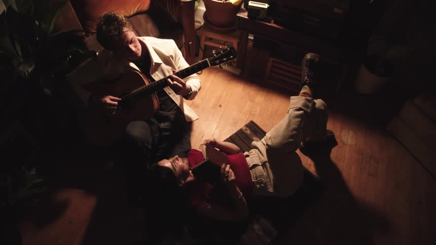 Man plays guitar seated while woman lies on floor using smartphone in warmly lit room with plants