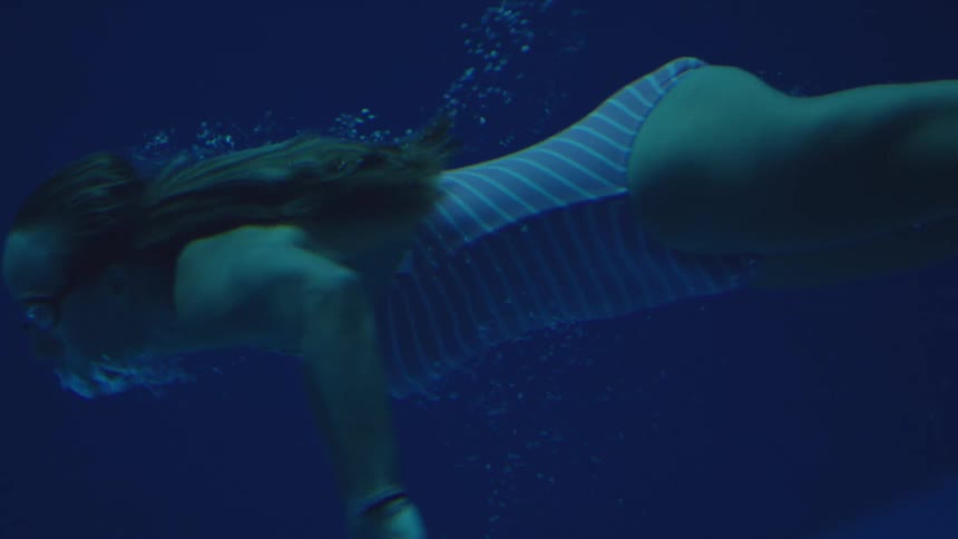Woman swims in horizontal position wearing striped swimsuit with loose hair in dark underwater setting