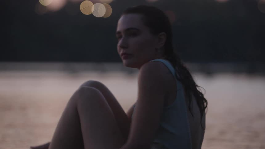 A woman with wet hair sits by calm water at dusk looking pensive