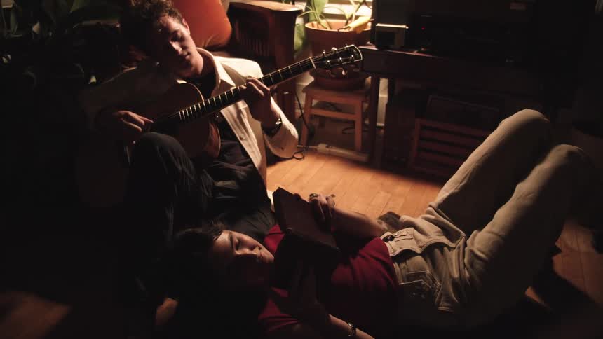 A man strums an acoustic guitar while a woman lies on the floor reading in warm lamplight both looking relaxed and cozy in a livedin room
