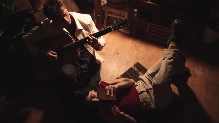 Man plays guitar while a woman lies beside him reading a book