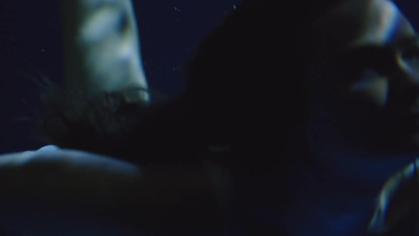 A woman swims underwater with hair streaming behind her her face in blue shadow with a focused neutral expression
