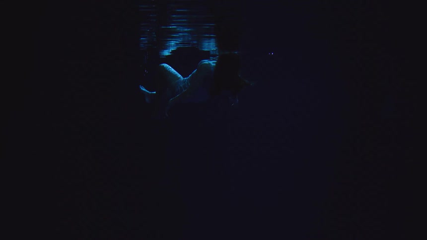 A dark underwater shot shows a human figure floating downward with their arms extended towards the waters surface which reflects bright fragmented blue light onto the figure
