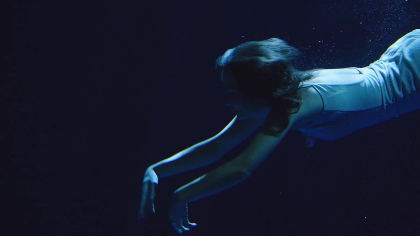 A woman in a pale blue outfit glides horizontally through the dark blue water with her arms extended as small bubbles rise past her creating a dramatic and moody feeling