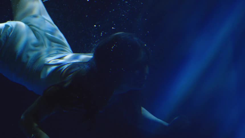 Woman submerged moves forward beneath blue beams of light in deep water