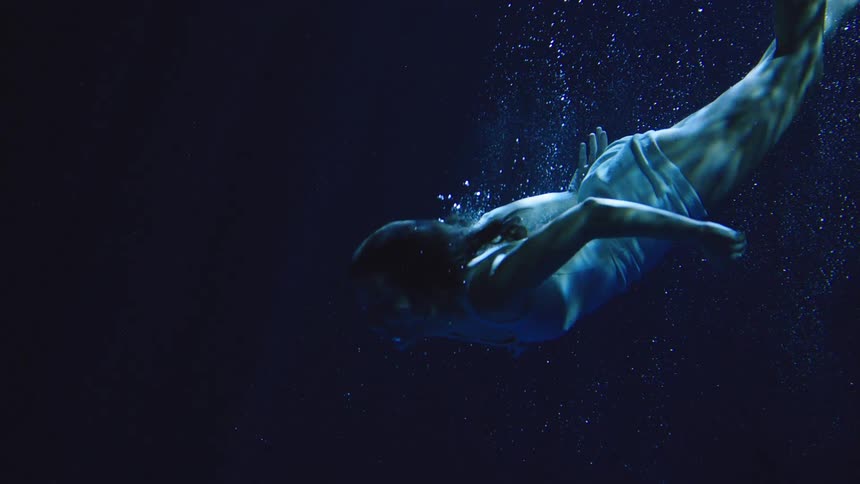 Person submerged middive wearing light outfit surrounded by bubbles in blue aquatic setting