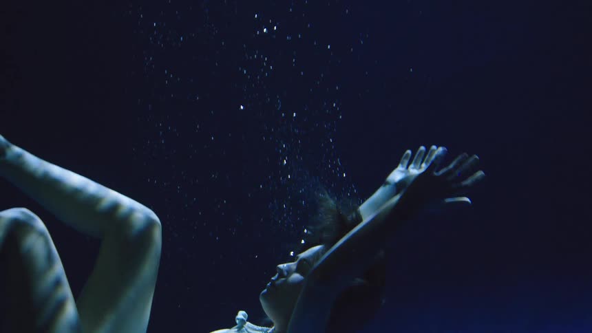 Woman underwater floats in a fetal position illuminated by deep blue light