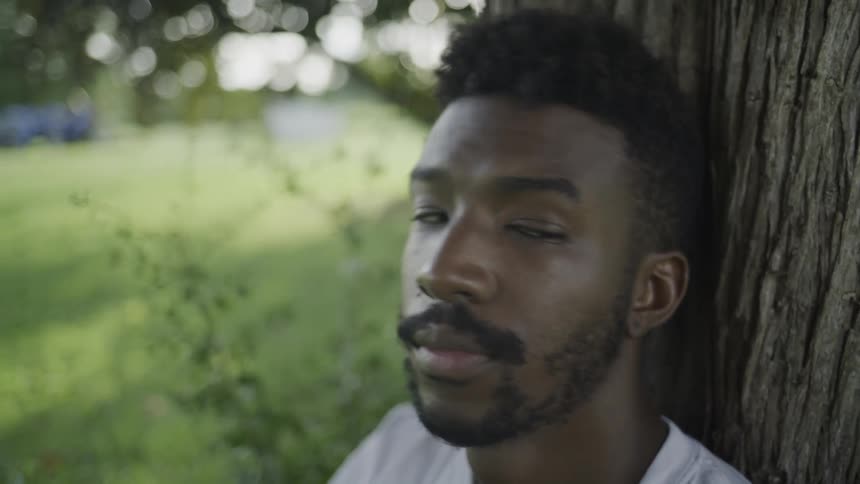 A close view of a man with short dark hair and a beard wearing a white shirt as he leans against the rough bark of a tree and looks at the camera in a green outdoor setting