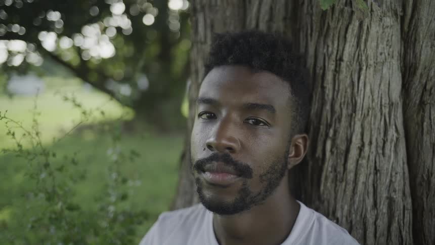 Black man wearing a white shirt sitting against a tree trunk outdoors during day time