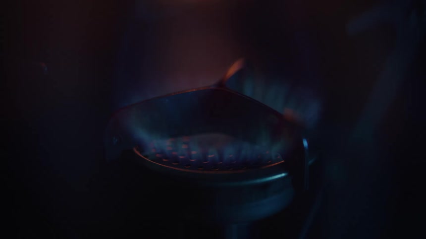 The video is an extreme closeup of a dark burner or stove element the flame is just beginning to ignite creating a small pale blue glow within the burners metal grating