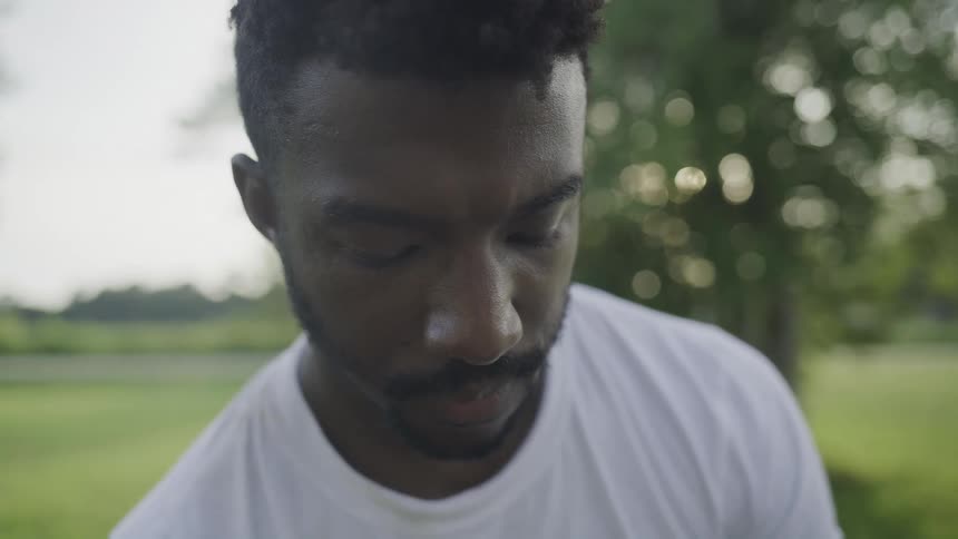 A young black male with a mustache stands on a sunlit grassland at midday wearing a white tshirt as the open field stretches around him under a clear blue sky
