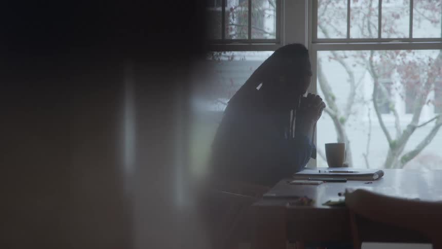 Black man sitting on a chair in front of a table frowning and praying indoors during day time