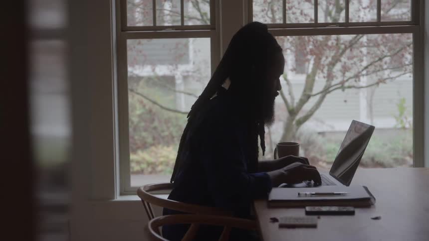 A man works on a laptop at a wooden table by a window