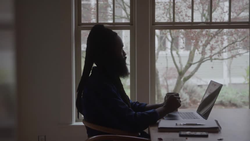 A man works at a desk with a laptop and mug near a bright window