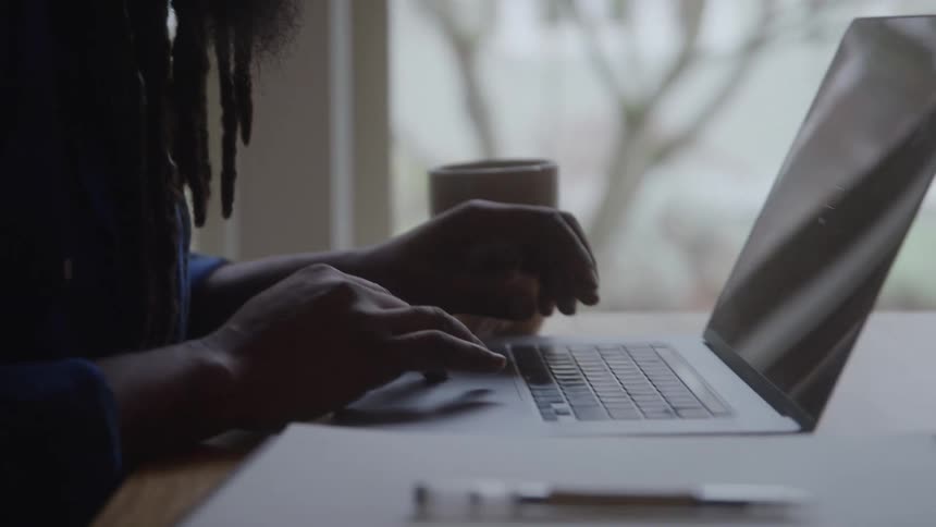 A young black male with long hair and a full beard types on his laptop at a desk a cup of coffee resting beside him as midday light filters into the workspace