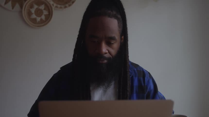 A man with long dark dreadlocks tied up in a ponytail wears a blue plaid shirt and focuses intently on a silver laptop in a brightly lit room with decorative woven baskets on the wall