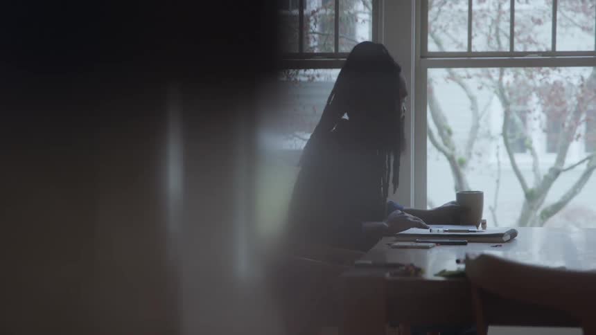 A young man with long braided hair and a full beard sits behind a desk closes his laptop lid and picks up a cup of coffee bringing it to his lips as midday light filters into the room