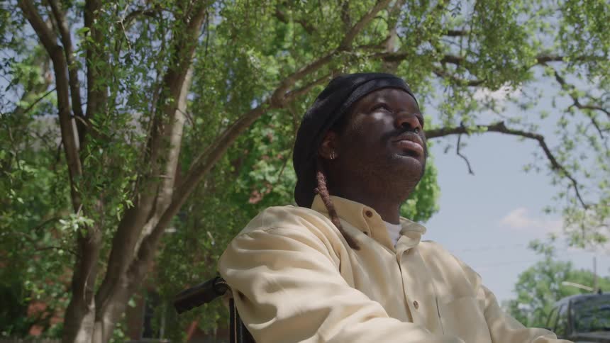 A young black male wearing a yellow shirt and black hat sits in a wheelchair under a tree in the woods on a sunny day staring at the sky in sadness