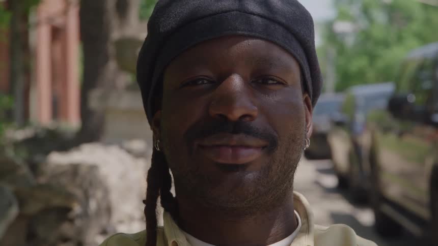 A close view shows a smiling man with dreadlocks wearing a black beret and a light colored shirt opening his eyes and looking directly at the camera on a sunny urban street