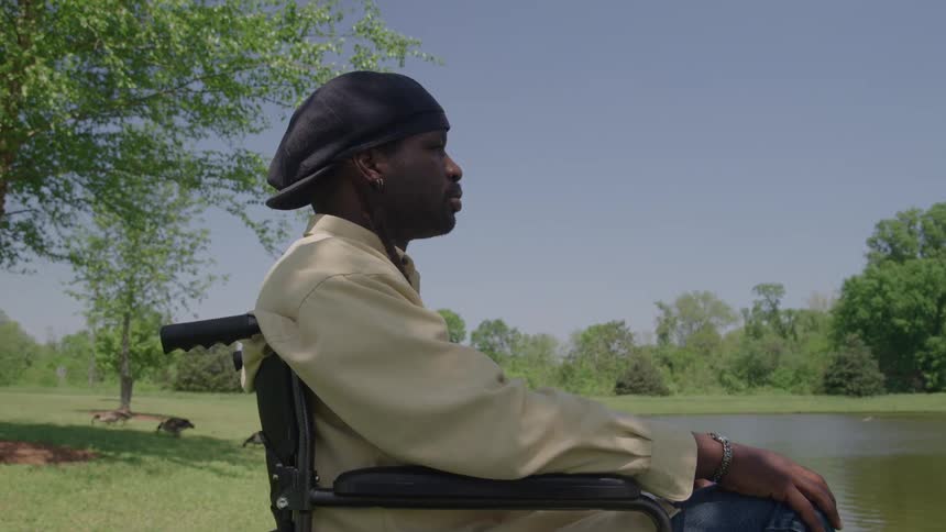 A black man in a wheelchair wearing a beret sits by a lakeshore under clear skies appearing calm and contemplative