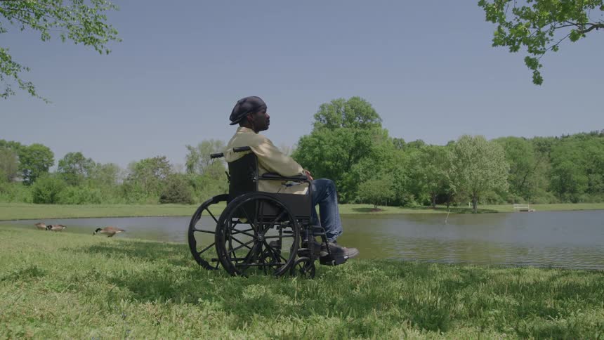 A man wearing a black hat a light yellow shirt and blue jeans sits in a wheelchair looking out over a calm lake surrounded by green trees under a clear blue sky with a couple of geese grazing nearby