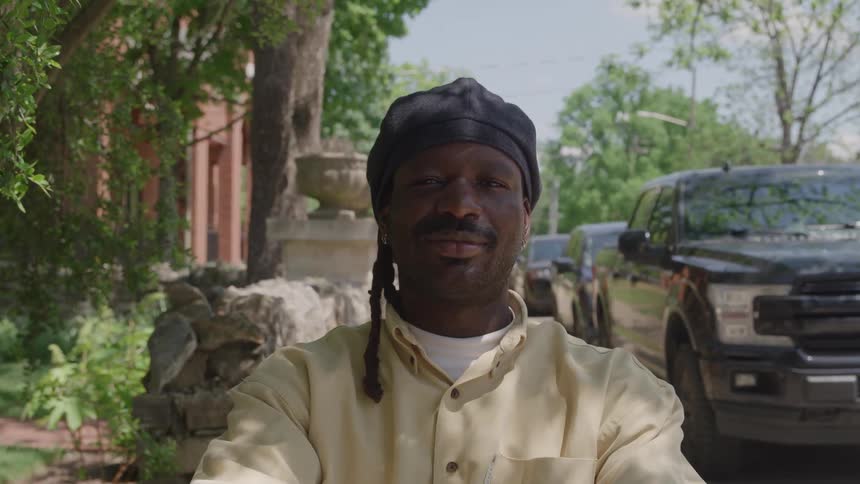 A man in a black cap and yellow shirt looks calmly at the camera on a leafy neighborhood street with parked vehicles