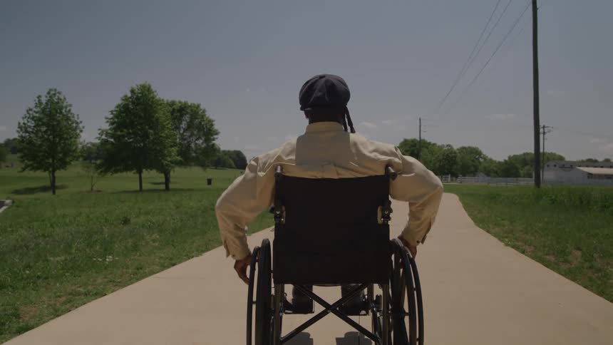 Black man wearing a hat rolling on a wheelchair on a suburban area surrounded with trees and vegetation outdoors during day time