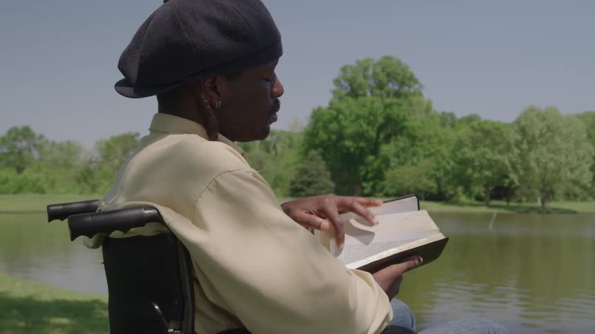 A man with short dreadlocks wearing a black cap and light yellow shirt sits in a wheelchair reading a book while relaxing by a green lake on a sunny day
