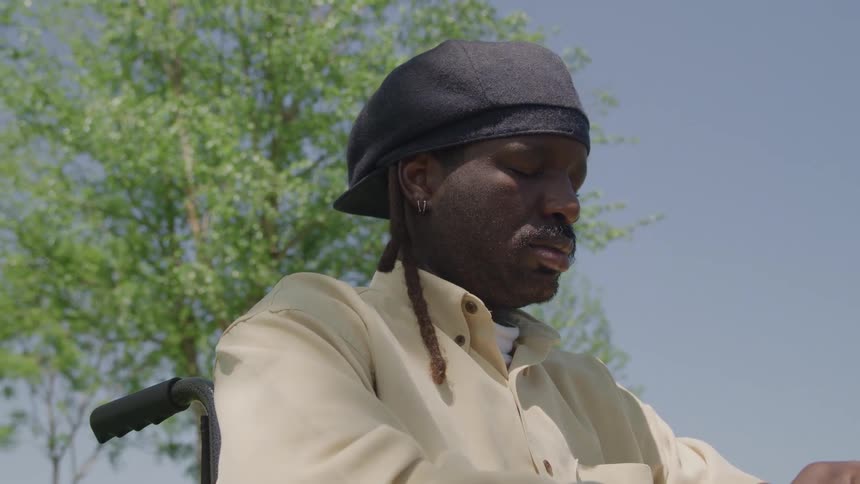 A young black male sits in a wheelchair beneath a tree on a sunny day wearing a black hat as dappled sunlight filters through the leaves casting soft shadows across the grass