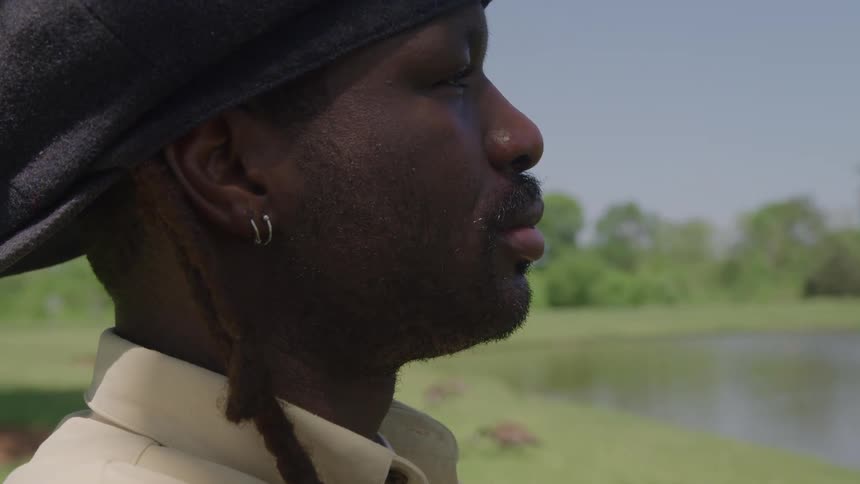 Black man wearing a black bonnet and a white shirt looking around on a natural setting surrounded by grass and trees in front of a body of water outdoors during day time