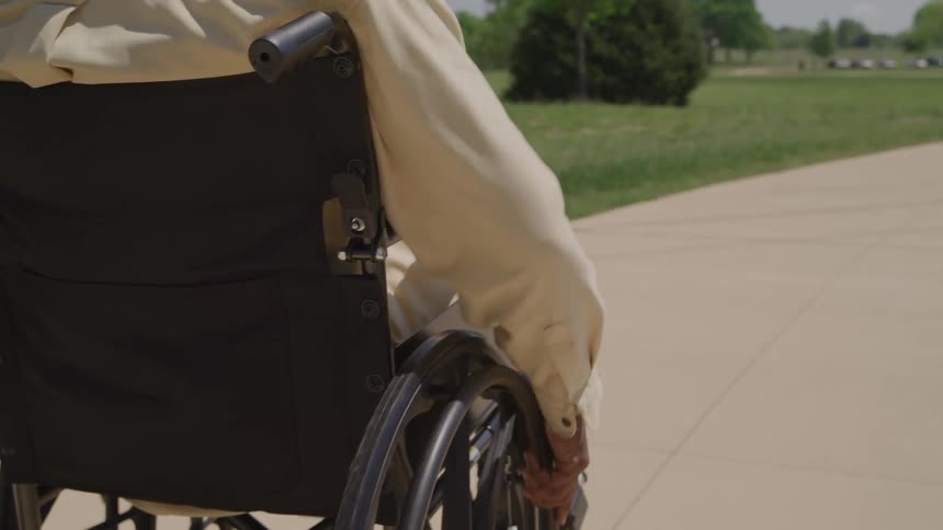The torso and arms of a man wearing a light colored shirt are seen from behind as he propels himself forward in a black wheelchair along a paved path in a sunny park