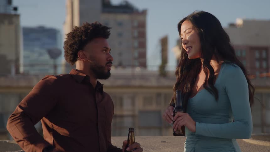 A young black male and an asian woman sit together on a rooftop during a sunny evening sharing drinks and enjoying each others company as warm light casts golden tones across the city skyline