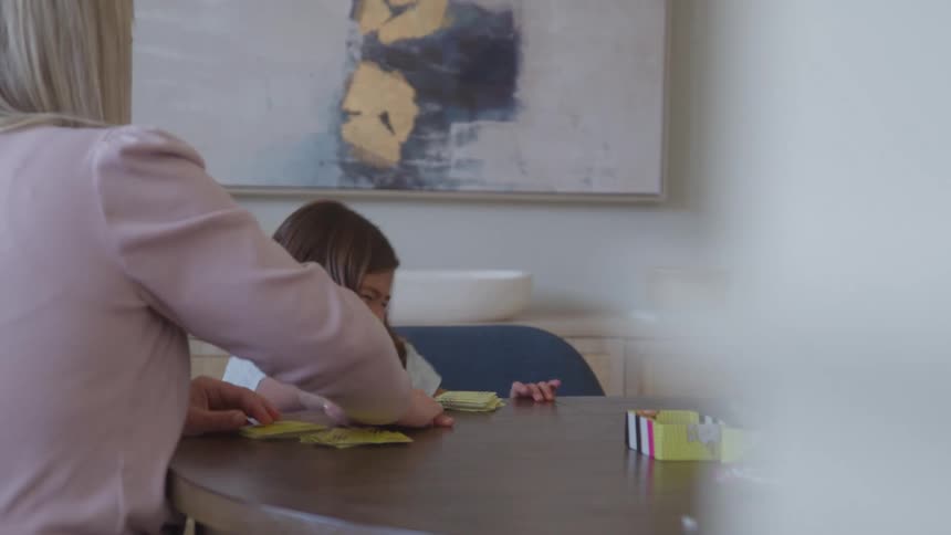 A happy young girl in a light blue shirt smiles while sitting at a wooden table playing a card game with an out of focus woman wearing a pink top in a bright dining area