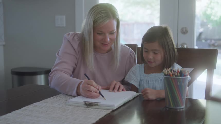A woman in a pink sweater draws a spiral with a black pencil in a sketchbook as a young girl in a light blue dress watches near colored pencils nearby on a wooden table