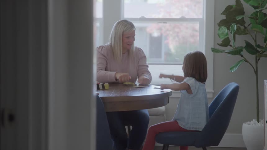 A woman in a pink sweater and a young girl in a light blue top sit at a round wooden table smiling and playing a card game in a brightly lit dining room