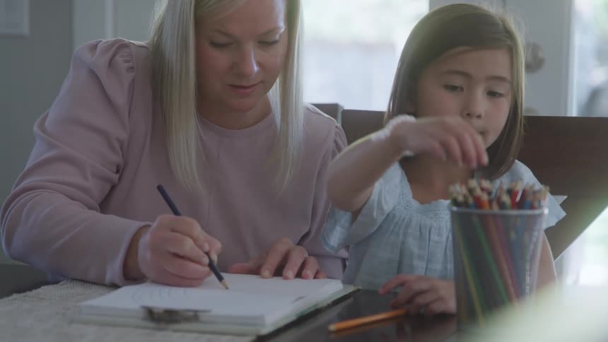 A woman in a pink sweater draws in a sketchbook with a pencil next to a young girl in a light blue dress who reaches for a red colored pencil from a container on a wooden table