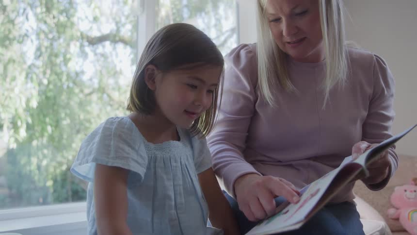 Woman reads a book with a girl sitting beside her in a bright room