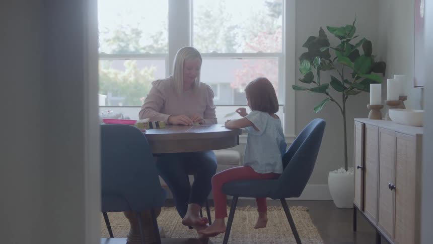A woman in a pink sweater and a young girl in a light blue shirt sit across a round wooden table from each other playing a game or activity in a bright dining room