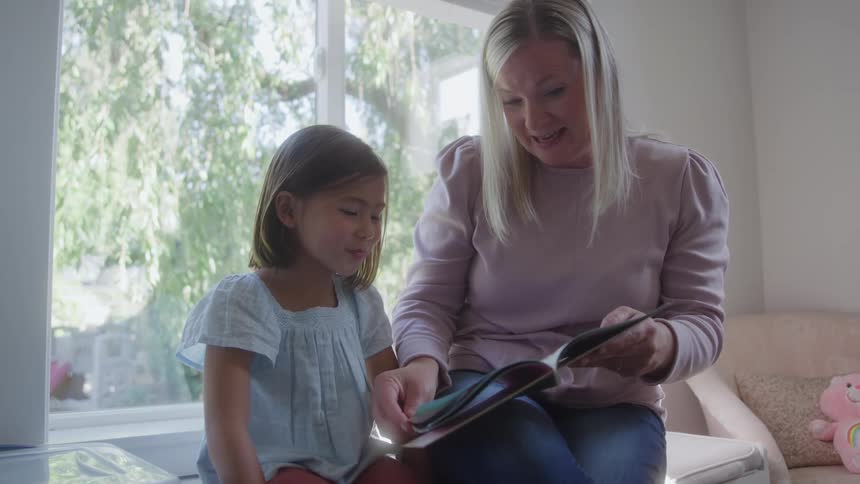 Mother reads book to daughter seated on bench by window with natural light and toys around